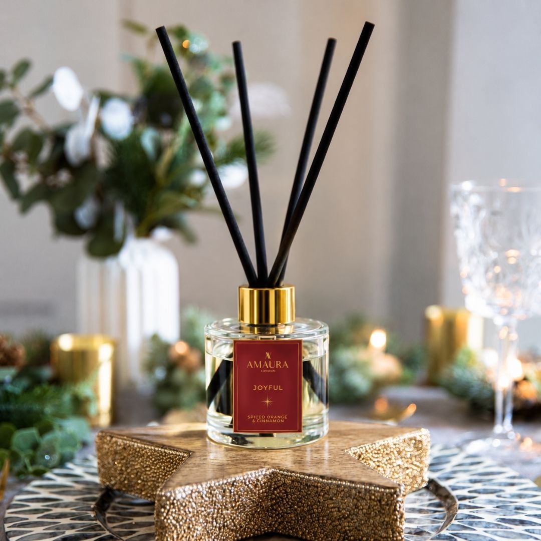 A reed diffuser bottle with black reeds, a gold cap, and a red label reading 'Amaura Joyful', Spiced Orange and Cinnamon scented reed diffuser